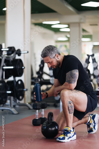 Fototapeta Naklejka Na Ścianę i Meble -  Sportsman with bottle of water and looking away while sitting on floor during break in fitness workout in gym