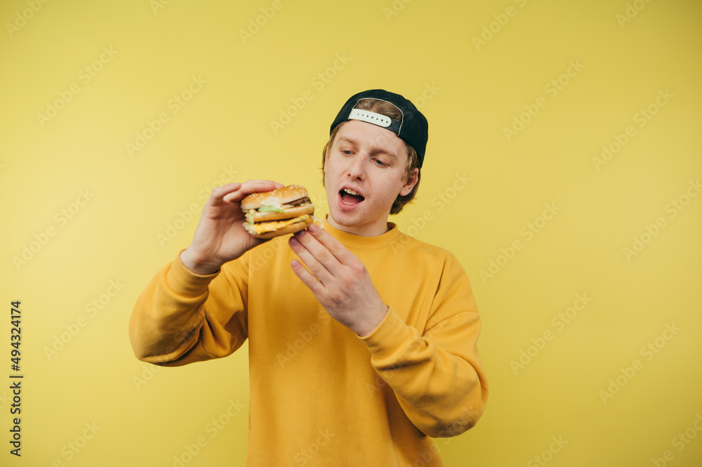 Handsome young man in a cap stands on a yellow background with a burger in his hands and looks at fast food with a hungry look. Handsome young man in a cap stands on a yellow background with a burger in his hands and looks at fast food with a hungry look.