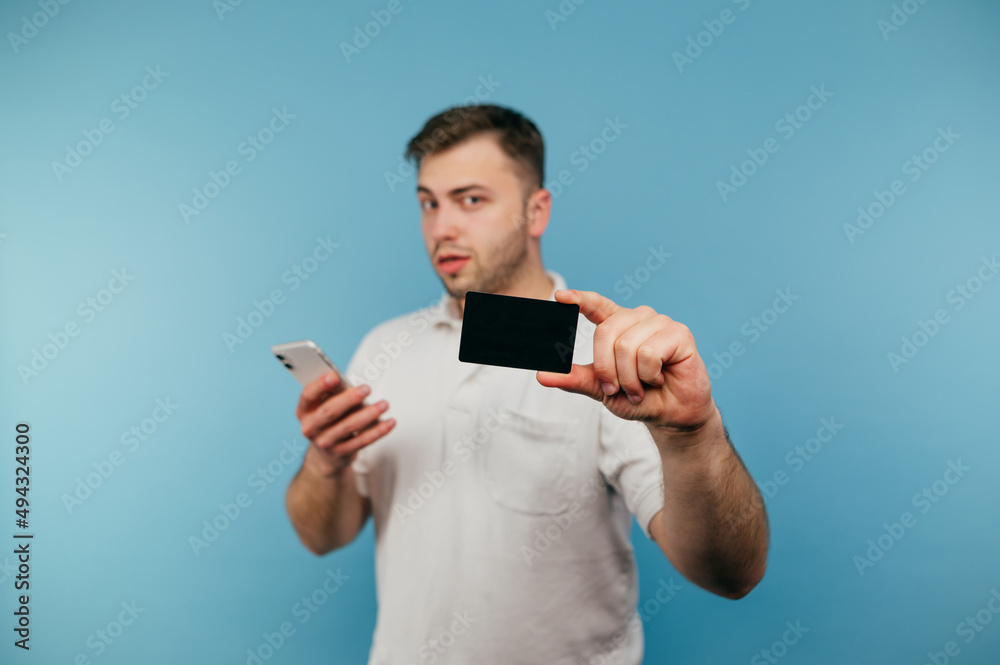 Handsome man in white t-shirt with smartphone in hands shows black bank card to camera isolated on blue background