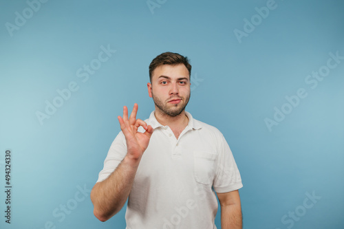 Adult serious man with bristles in a white T-shirt shows gesture ok to the camera and looks intently, isolated on blue background