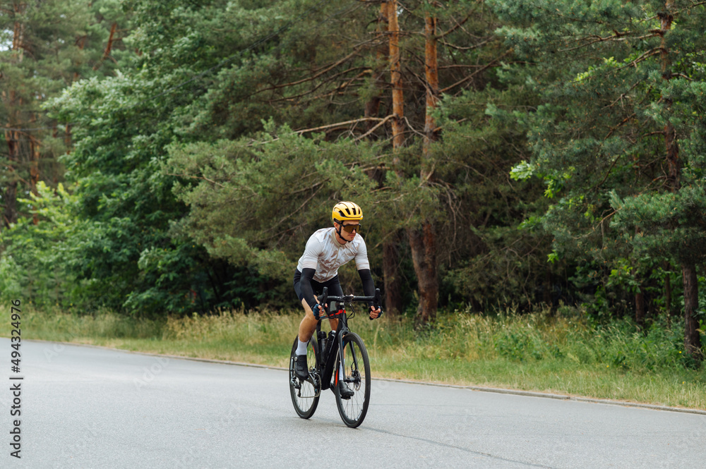 Athletic handsome cyclist in outfit trains out of town in the woods on asphalt road, rides fast forward, side view