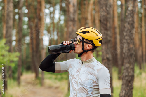 Handsome young man cyclist in helmet and goggles drinks water from a bottle on a background of pine forest and looks away.
