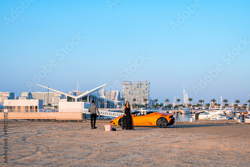 Couple with luxury supercar on sand with river and cityscape in the background