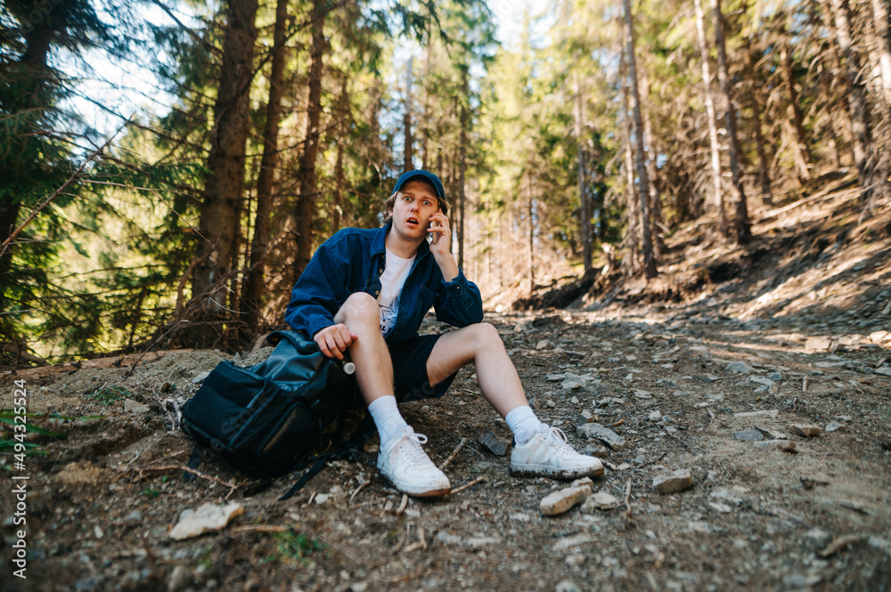 Shocked young male tourist sitting with a backpack, resting on a trail while climbing mountains, talking on the phone and looking at the camera with a surprised face