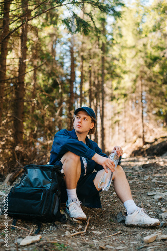 Handsome young man sitting in the woods with a backpack while hiking in the mountains with a bottle of water in his hands, looking away with a serious face.