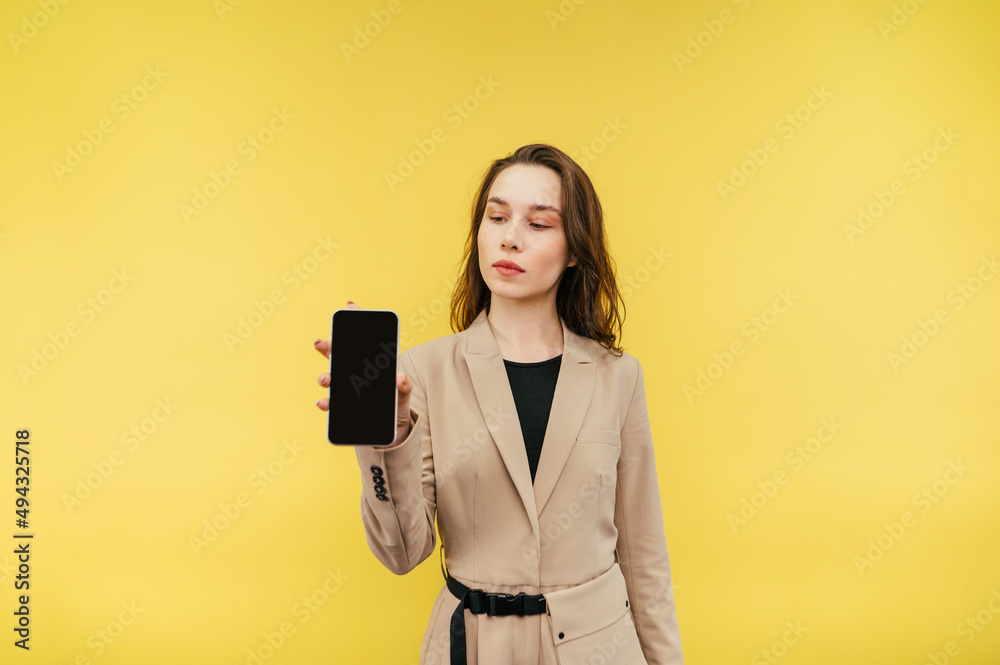 Beautiful woman in a suit with a smartphone in his hands stands on a yellow background and shows the camera screen with a black screen. Beautiful woman in a suit with a smartphone in his hands stands on a yellow background and shows the camera screen with a black screen.