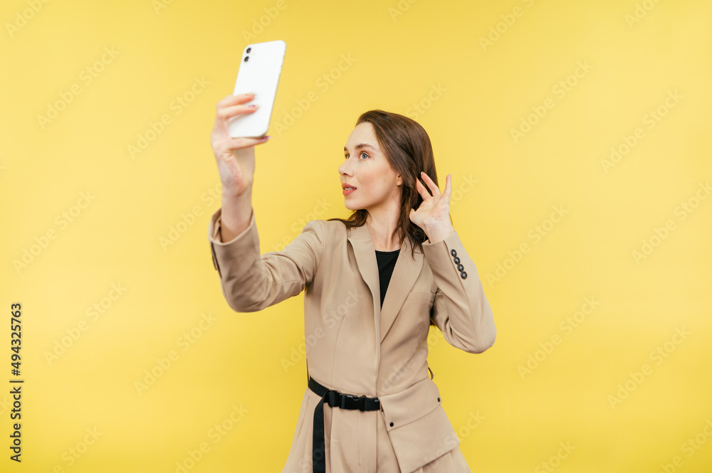Naklejka premium Beautiful woman in jacket making selfie on yellow background, posing on smartphone camera, isolated.