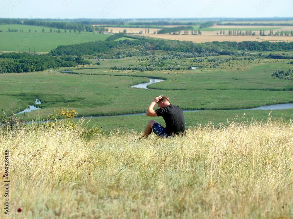 A man sitting on the edge of a cliff looks admiringly at the riverbed, amid green meadows. In the background you can see fields of wheat and trees, shot in sunny weather. A view of Ukrainian fields A man sitting on the edge of a cliff looks admiringly at the riverbed, amid green meadows. In the background you can see fields of wheat and trees, shot in sunny weather. A view of Ukrainian fields