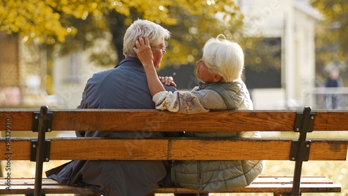 Aged Caucasian married couple sitting on the bench woman touching his husband's hair smiling medium shot from the back selective focus . High quality photo
