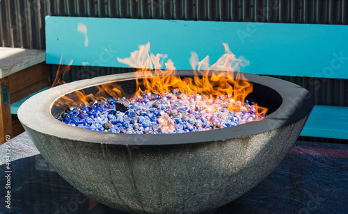 Circular fire pit with flames and blue rock lava in outdoor setting near benches providing warmth.