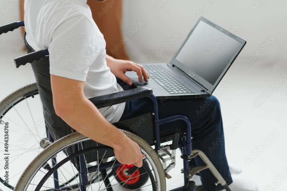 Obraz premium Portrait of smiling disabled male sitting in wheelchair and working on laptop from home. Young worker with special needs. Freelancer and people with disabilities concept.