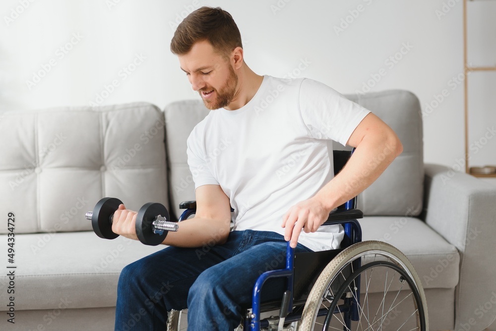 sportsman sitting in wheelchair and outstretching arms with dumbbells during rehabilitation exercise in modern medical center. Man sitting on wheelchair at home. sportsman sitting in wheelchair and outstretching arms with dumbbells during rehabilitation exercise in modern medical center. Man sitting on wheelchair at home.