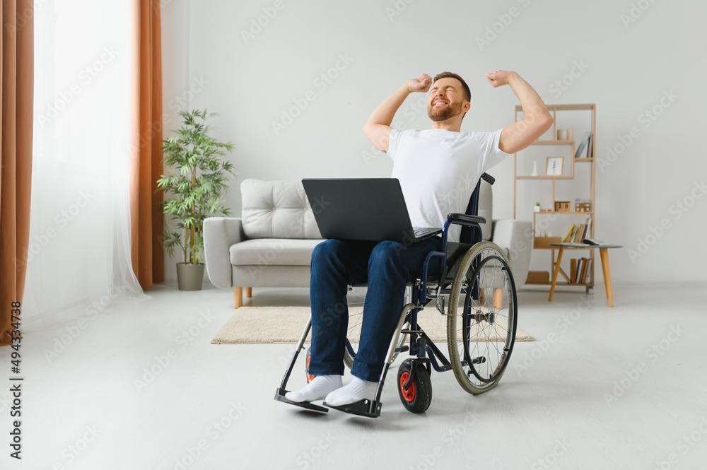 Portrait of smiling disabled male sitting in wheelchair and working on laptop from home. Young worker with special needs. Freelancer and people with disabilities concept. Portrait of smiling disabled male sitting in wheelchair and working on laptop from home. Young worker with special needs. Freelancer and people with disabilities concept.