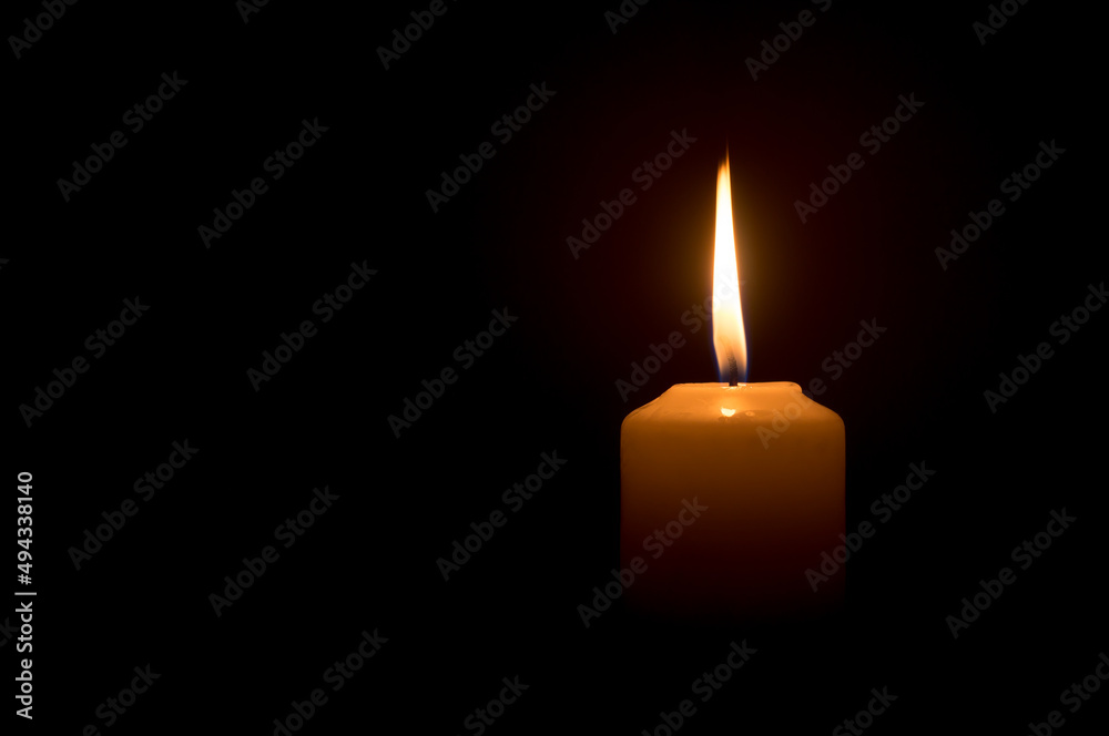 A single burning candle flame or light glowing on a white candle on black or dark background on table in church for Christmas, funeral or memorial service A single burning candle flame or light glowing on a white candle on black or dark background on table in church for Christmas, funeral or memorial service