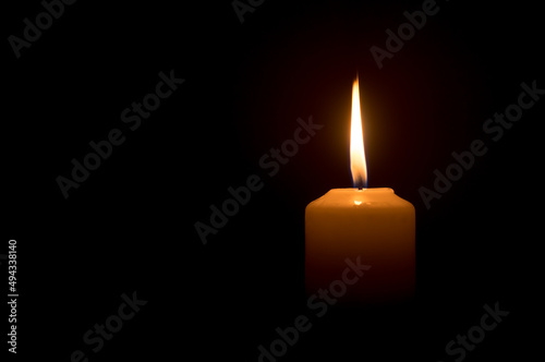 A single burning candle flame or light glowing on a white candle on black or dark background on table in church for Christmas, funeral or memorial service