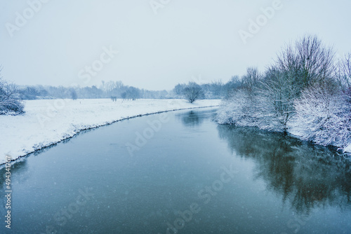 Beautiful view of a river flowing in a snow-land with leafless trees on its banks against cloudy sky photo