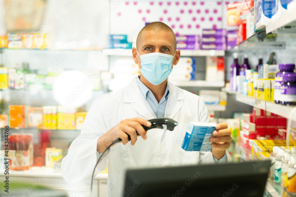 Male pharmacist in lab coat and face mask standing at counter in drugstore and using barcode scanner to sale drug package.