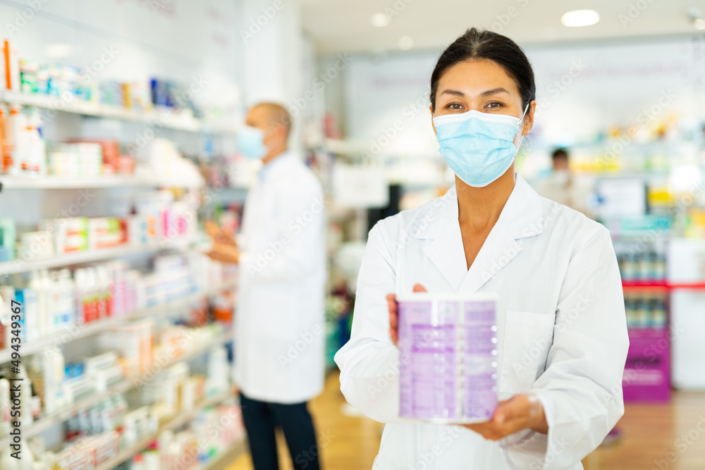 Asian female doctor in face mask standing in salesroom of chemist shop and holding can with infant formula in hands. Her colleague working in background.