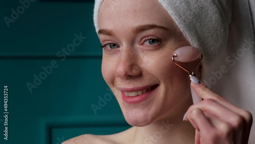 Close-up portrait of a woman using jade roller tool for facial massage after bath, smiling with pleasure. Dermatology, cosmetology. Healthy lifestyle photo