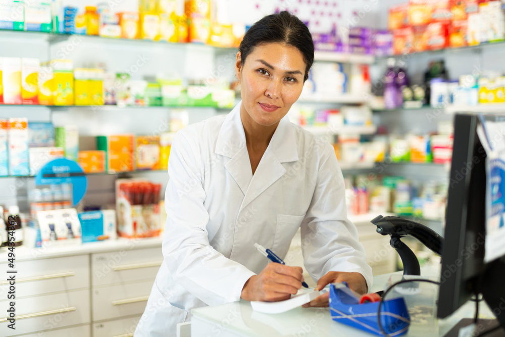Oriental female pharmacist standing at counter in chemist shop and writing recipe.