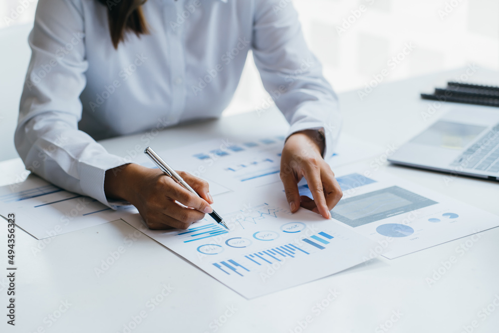 Asian woman is pointing at the report graph and has laptop on the front. Maybe preparing data for marketing report. window light Asian woman is pointing at the report graph and has laptop on the front. Maybe preparing data for marketing report. window light