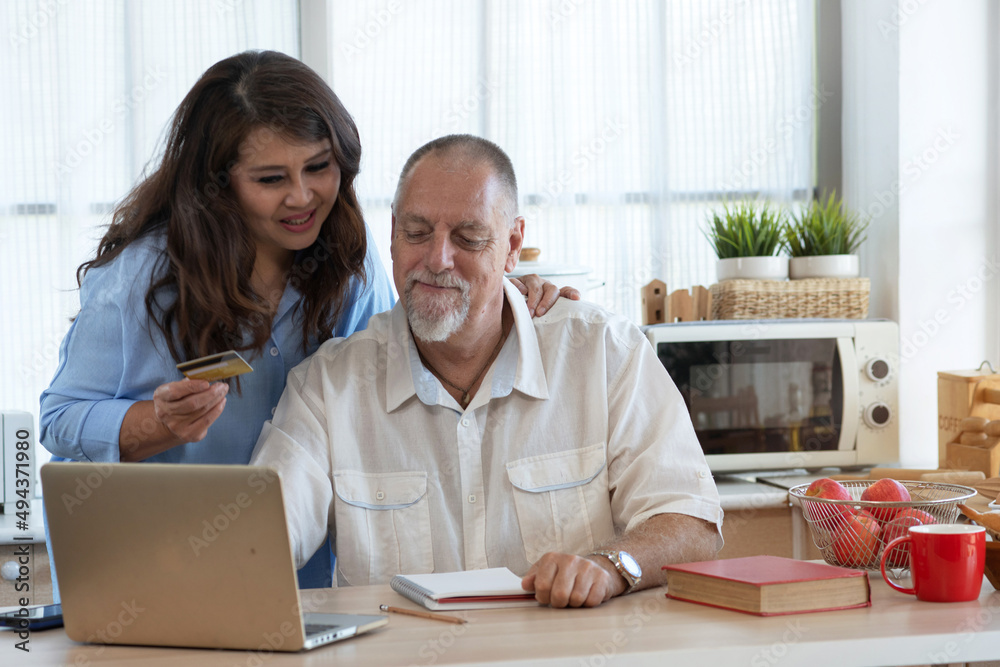 Mature middle-aged couple using credit card for online shopping, buying at home, using laptop computer in kitchen at home, woman holding credit card Mature middle-aged couple using credit card for online shopping, buying at home, using laptop computer in kitchen at home, woman holding credit card