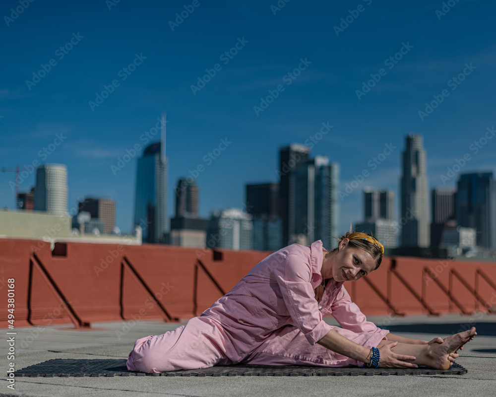 Young blonde woman in pink coveralls and barefoot practices yoga on a downtown Los Angeles Roof top with the LA skyline behind her