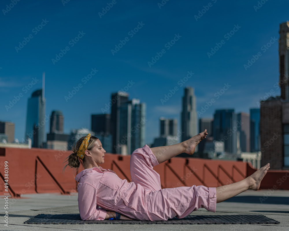 Young blonde woman in pink coveralls and barefoot practices yoga on a downtown Los Angeles Roof top with the LA skyline behind her Young blonde woman in pink coveralls and barefoot practices yoga on a downtown Los Angeles Roof top with the LA skyline behind her