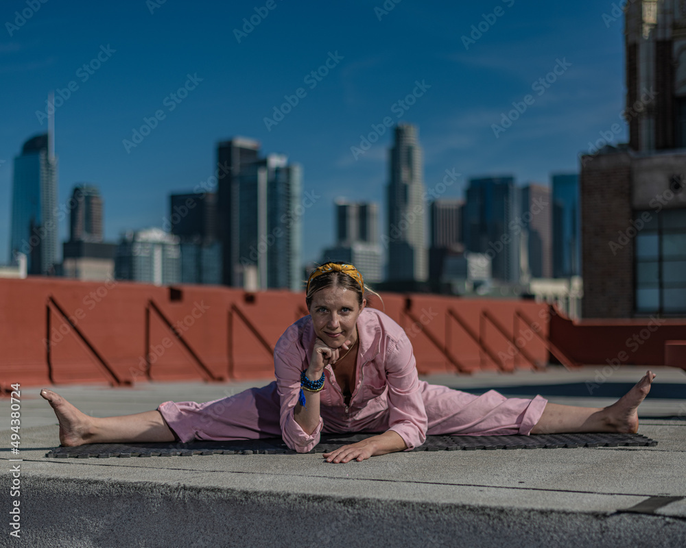 Young blonde woman in pink coveralls and barefoot practices yoga on a downtown Los Angeles Roof top with the LA skyline behind her Young blonde woman in pink coveralls and barefoot practices yoga on a downtown Los Angeles Roof top with the LA skyline behind her