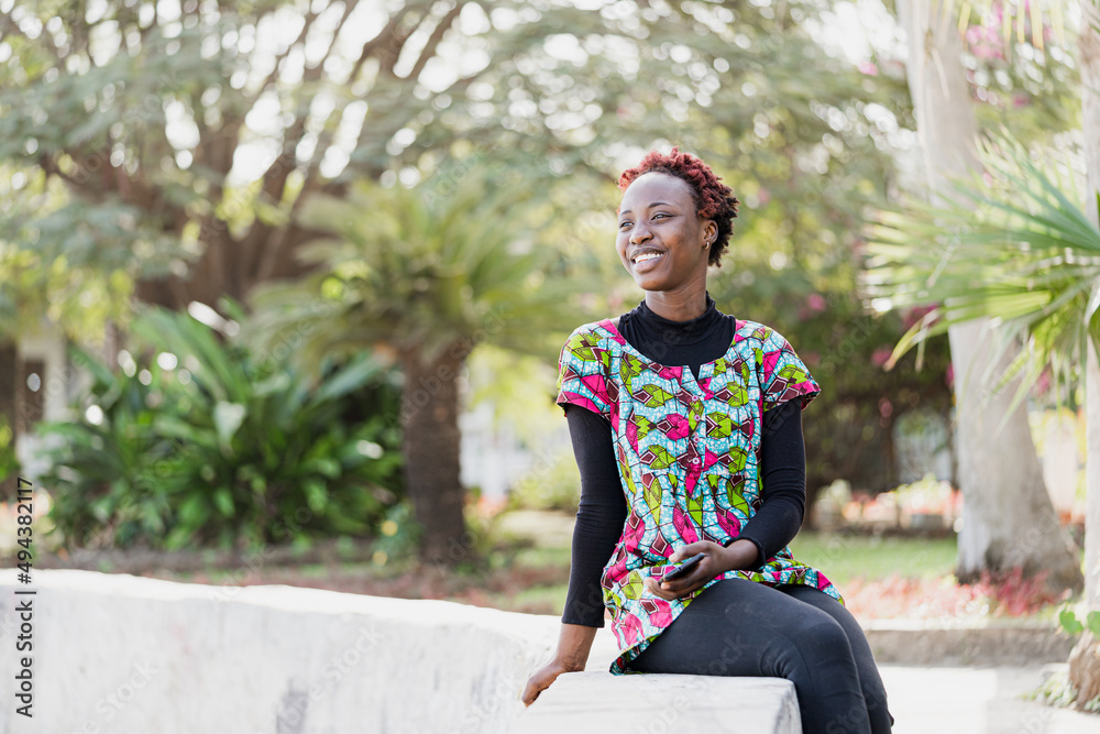 Modern African teenager in casual wear posing on a low wall in a green garden with her smartphone in her hand, youth and happiness concept