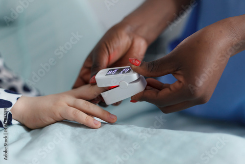 Close up of pediatric clinic nurse measuring sick child oxygen levels while in patient recovery ward. Hospital staff checking sick little girl oxygen level while laying relaxed on patient medical bed.