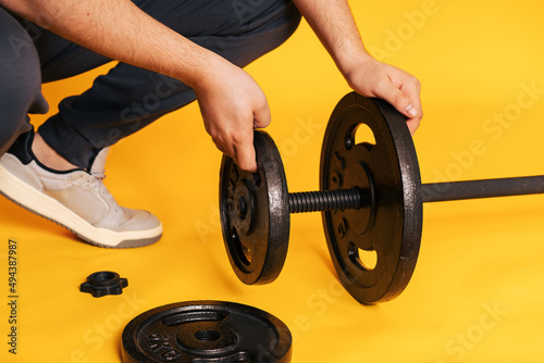 Man puts pancakes on barbell with his hands against yellow background.