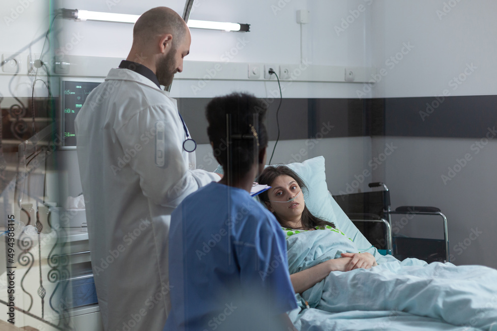 Medical doctor and nurse doing consult in private ward to recovering woman in hospital bed after surgical intervention. Patient with nasal cannula looking at health care professionals. Medical doctor and nurse doing consult in private ward to recovering woman in hospital bed after surgical intervention. Patient with nasal cannula looking at health care professionals.