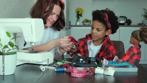 Caucasian mom and an African-American daughter sew clothes together for a doll at home on a sewing machine.Diverse people.Time together.Mother's day.Slow motion.