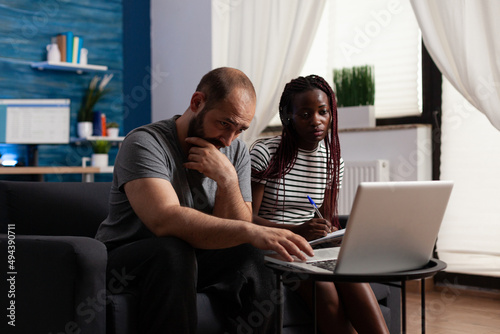 Worried multiethnic couple looking at laptop screen while african american woman taking notes. Stressed young adults working together to solve wrong banking bills using online accounting