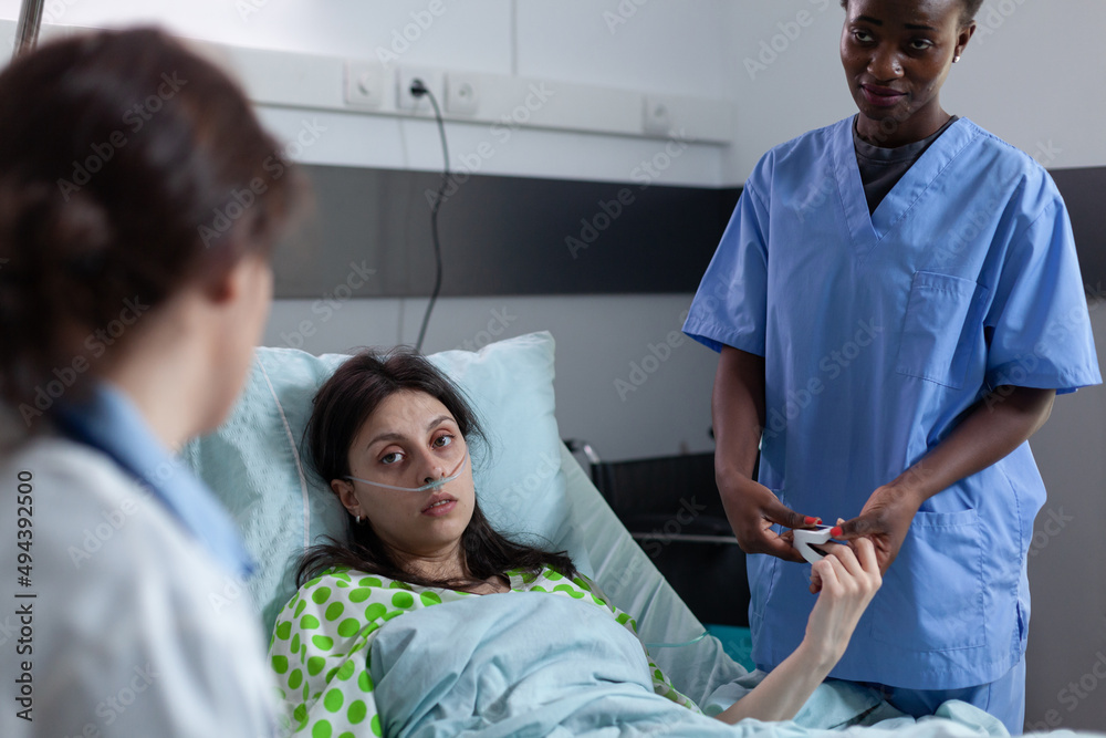 Over shoulder view of woman with low spo2 saturation recieving oxygen recovering in hospital bed looking at doctor providing medical care. Nurse attending patient with nasal cannula in hospital ward. Over shoulder view of woman with low spo2 saturation recieving oxygen recovering in hospital bed looking at doctor providing medical care. Nurse attending patient with nasal cannula in hospital ward.