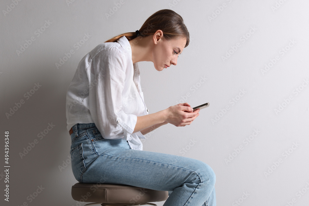 Woman with bad posture using smartphone while sitting on stool against light grey background - obrazy, fototapety, plakaty 