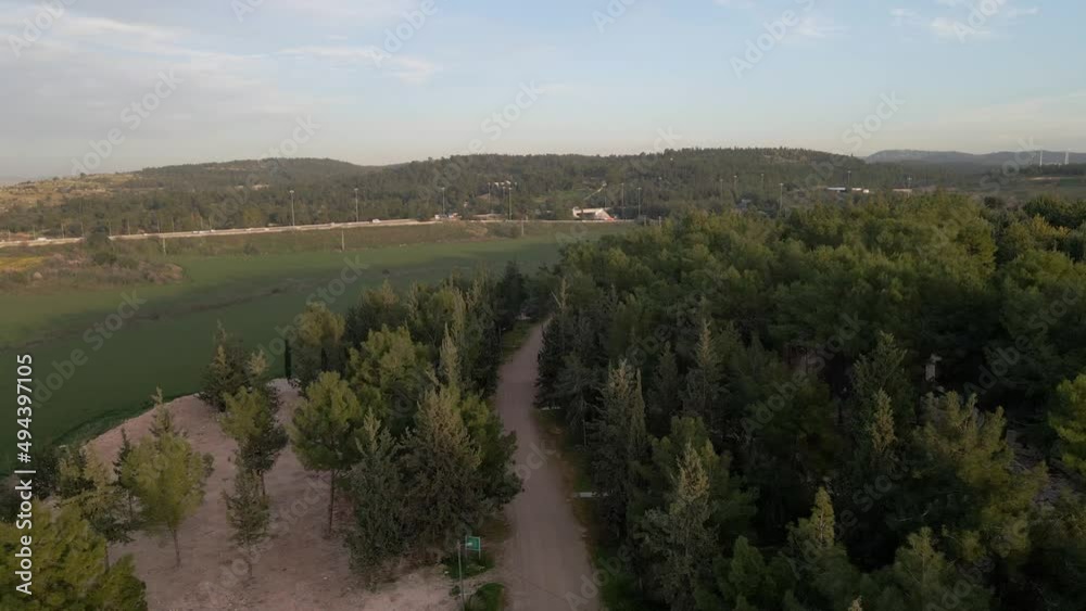 Aerial drone shot over a green forest in Latrun in Israel at sunset over a hilly terrain with the view of a busy highway in the background.