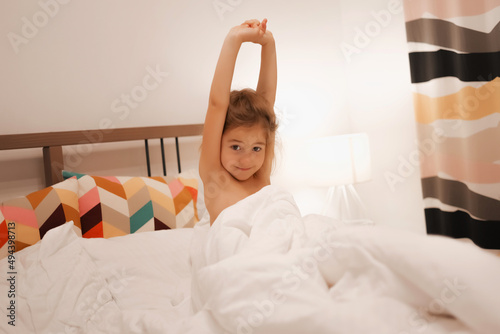 A European girl has woken up and is lying in bed with white pastel linen. The child has woken up, is pulling himself up and doing exercises photo