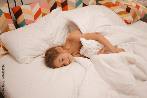 A European girl has woken up and is lying in bed with white pastel linen. Early in the morning, the child wakes up in the garden or school. Bright interior, striped pillows