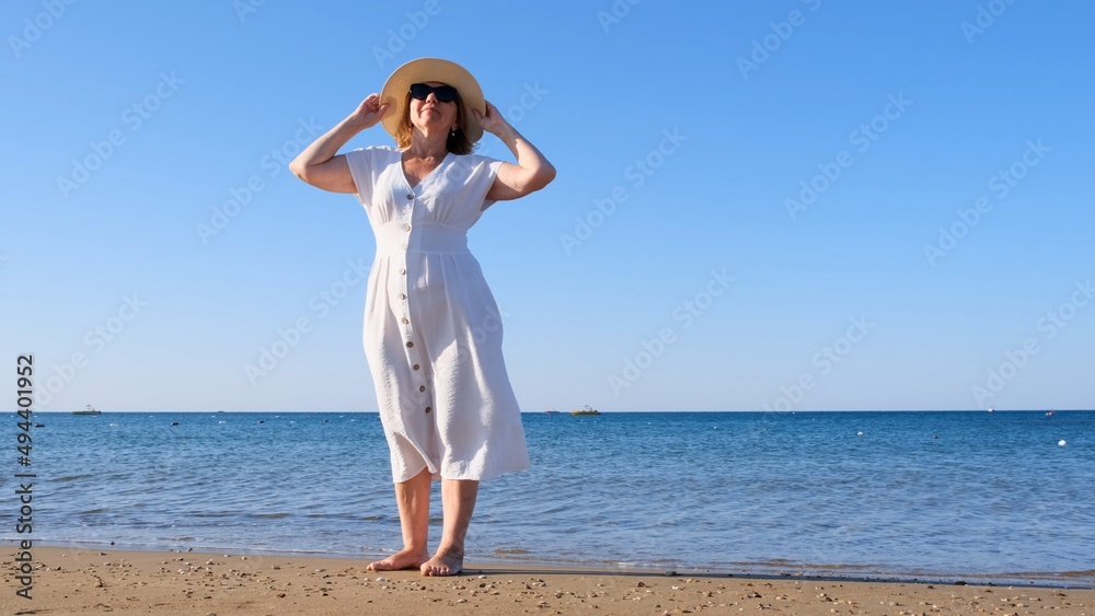 mature woman in a straw hat and white dress enjoying the sun walking along the blue sea coast on a sunny summer day, enjoying freedom and relaxation. The concept of a senior citizen everyday life mature woman in a straw hat and white dress enjoying the sun walking along the blue sea coast on a sunny summer day, enjoying freedom and relaxation. The concept of a senior citizen everyday life
