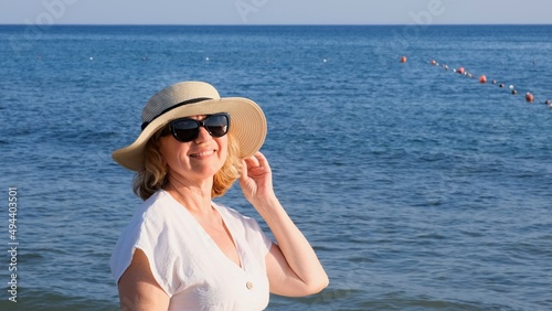 Happy mature woman 50 years old dressed in white dress, straw beach and sunglasses on the beach near the seashore. Vacation concept, relaxation, retirement age