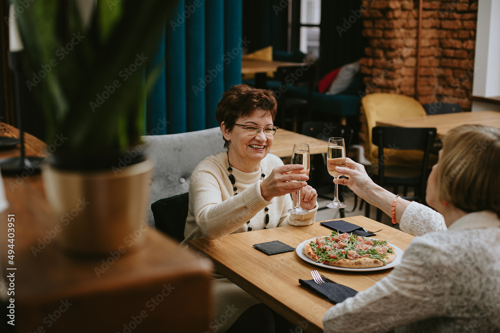 Two smiling elderly women with dark and fair hair clinking glasses of champagne sitting at table with pizza celebrating. Two smiling elderly women with dark and fair hair clinking glasses of champagne sitting at table with pizza celebrating.