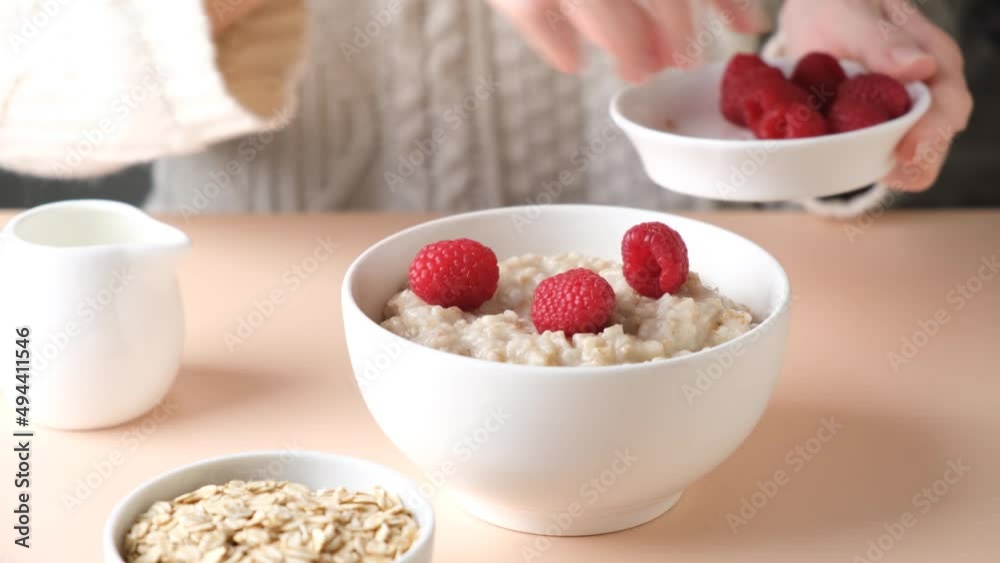 Oatmeal porridge with raspberries for breakfast. Woman adding fresh raspberries into cooked oatmeal bowl Oatmeal porridge with raspberries for breakfast. Woman adding fresh raspberries into cooked oatmeal bowl