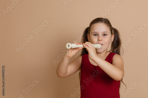 Girl in red dress play melody on flute with pleasure, blowing air into duct, beige background. Learn to play woodwind musical instrument. Flute and children is concept of music education development.