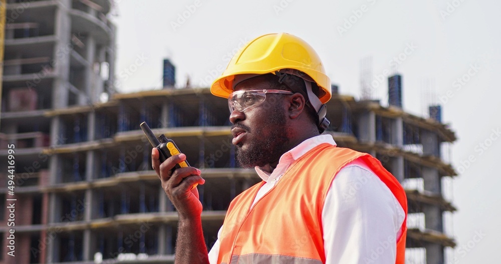 Builder in helmet is standing in front of the construction site, talking by walkie  talkie control and communicate with worker on construction site. Contractor  man using radio operation in industry. Stock Photo