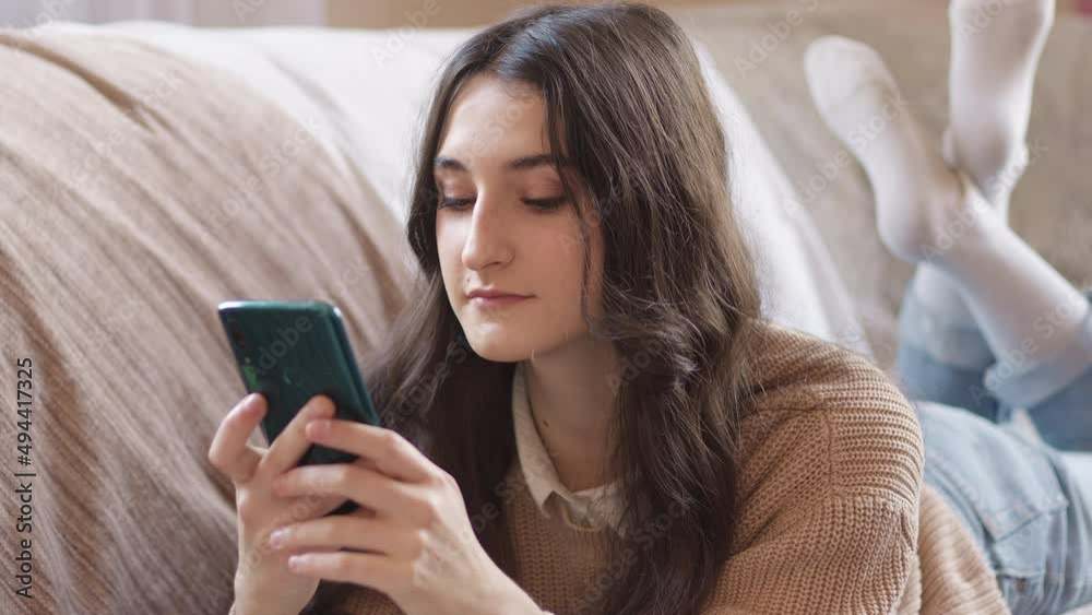 Smiling girl chatting in social network using phone while lying on sofa at home. Smiling girl chatting in social network using phone while lying on sofa at home.