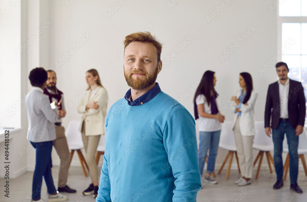 Portrait of confident young man against background of his colleagues from business team. Caucasian bearded male leader or company manager in casual clothes smiling looking at camera. Business concept.