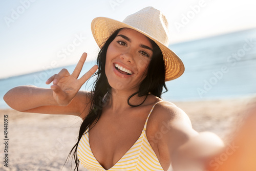 people, summer and swimwear concept - happy smiling young woman in bikini swimsuit and straw hat taking selfie on beach and showing peace gesture