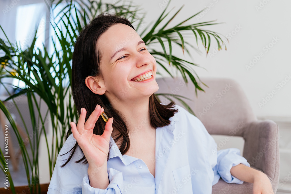 Smiling young woman with short hair holding pill omega capsule in hand at home. Girl taking medicine indoors Smiling young woman with short hair holding pill omega capsule in hand at home. Girl taking medicine indoors